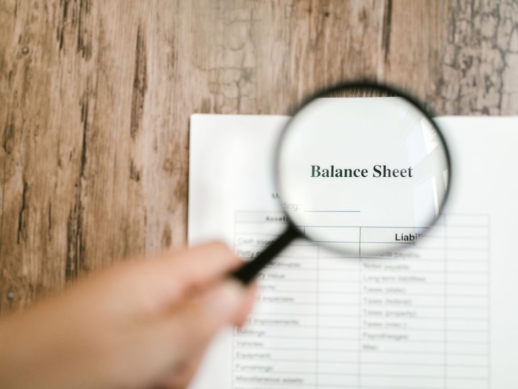 Close-up of a balance sheet document on wooden surface with a magnifying glass held by a hand