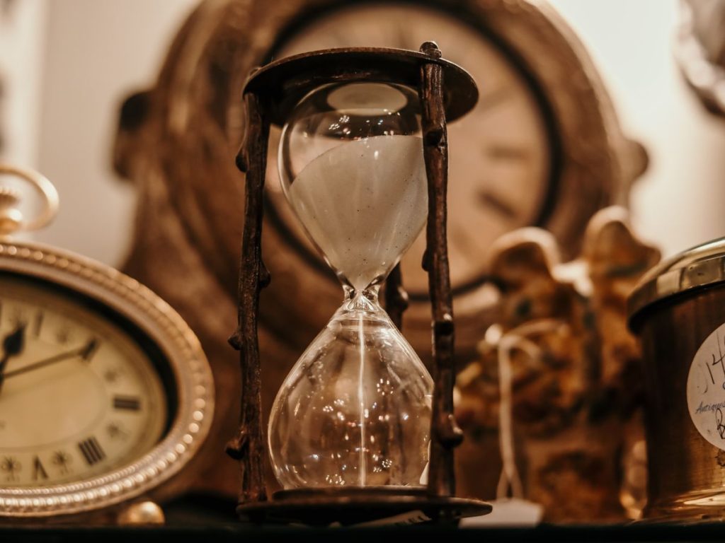 Close-up of a vintage hourglass surrounded by antique clocks in warm lighting