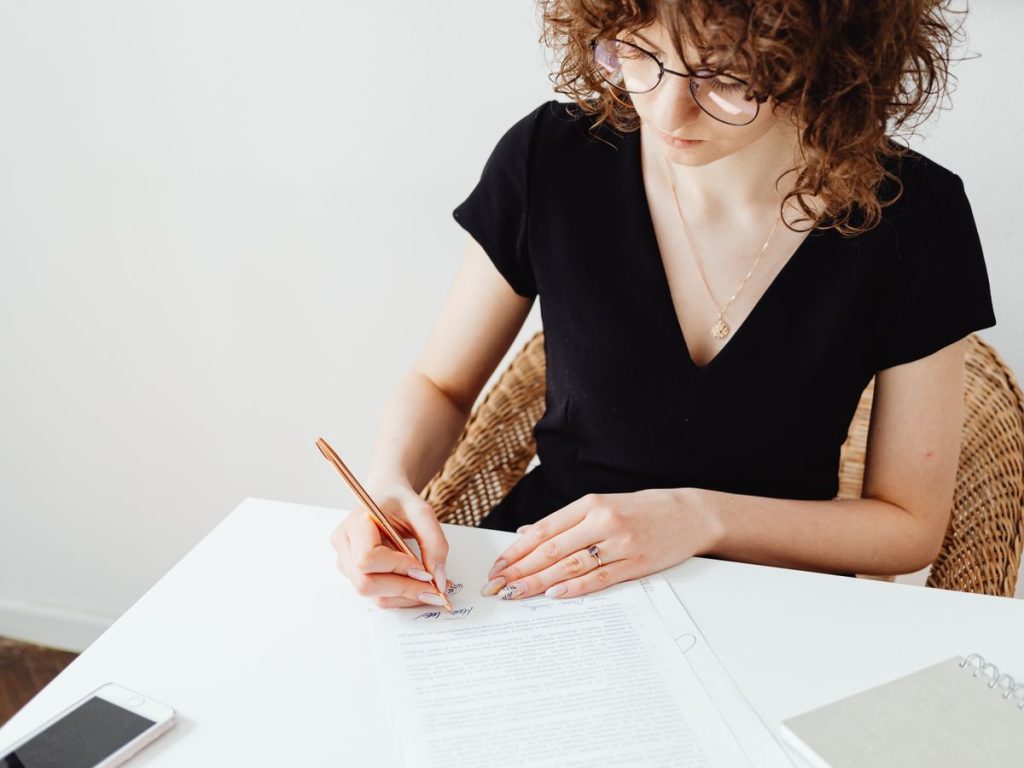 A woman in glasses and black top writing on documents at a white table, focusing intently