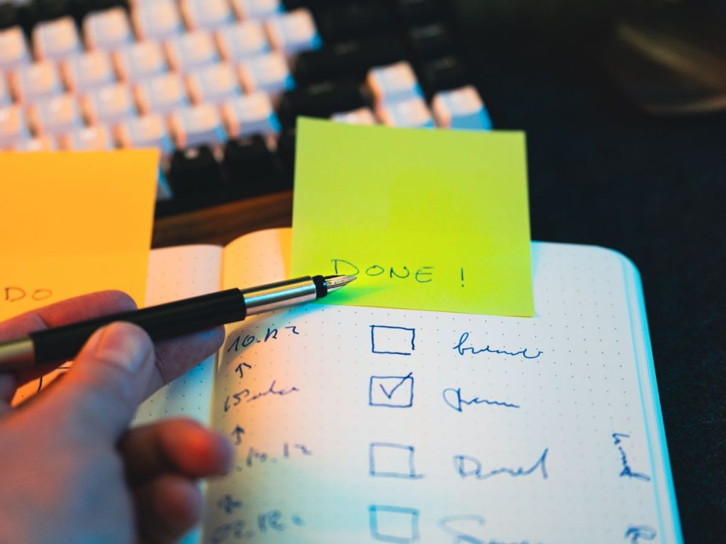 Close-up of hand holding pen over notebook checklist with sticky notes labeled 'DONE!'