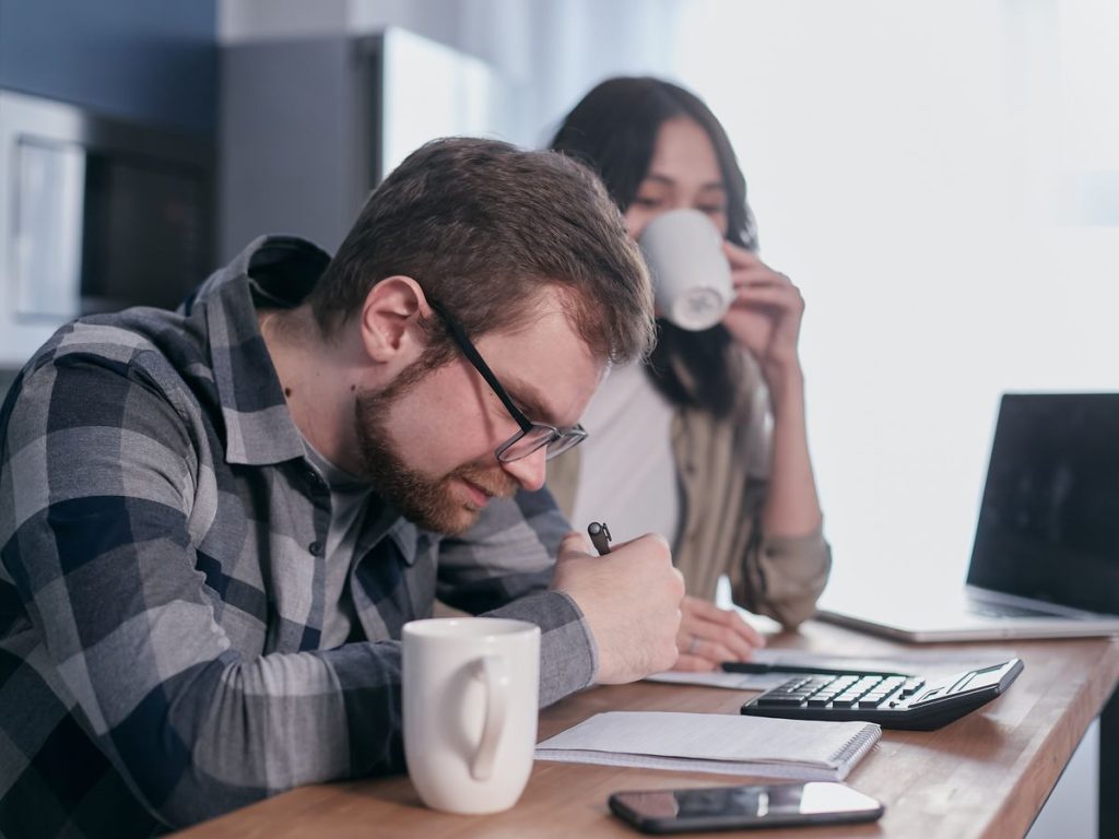 A couple sits at a kitchen table, reviewing finances with a laptop and calculator