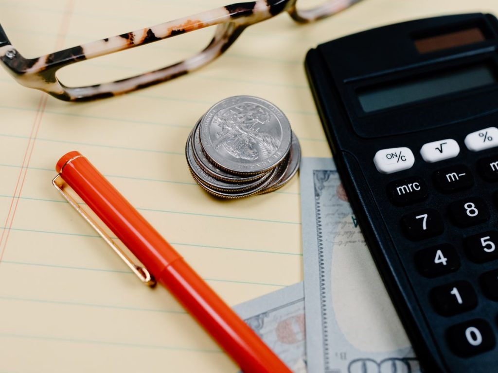 Close-up of a workspace with a calculator, coins, and glasses on a notepad