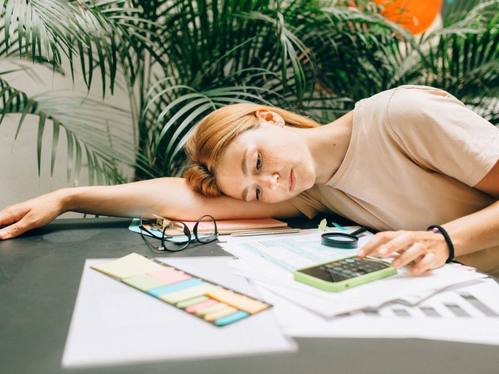 A tired woman calculating her taxes at a desk covered with papers and a smartphone