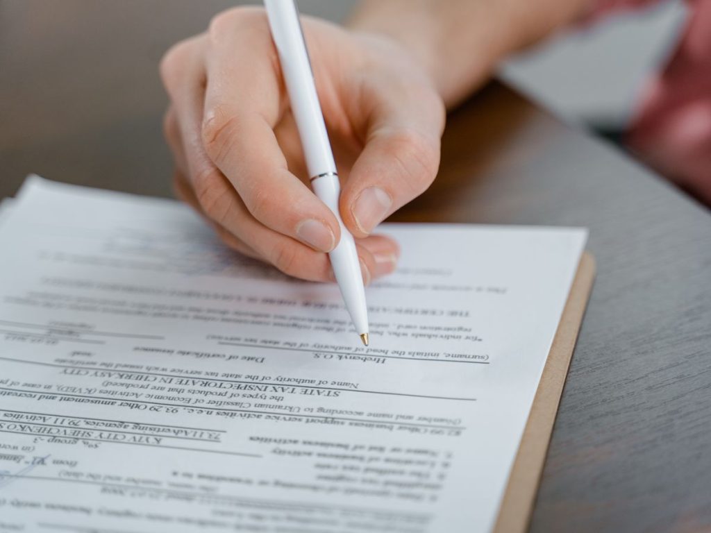 A hand holding a pen signing a document, close-up shot with focus on the paper