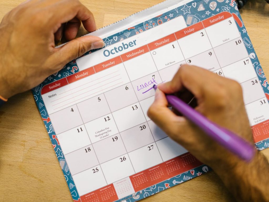 Close-up of a person writing a lunch reminder on an October calendar with a purple pen