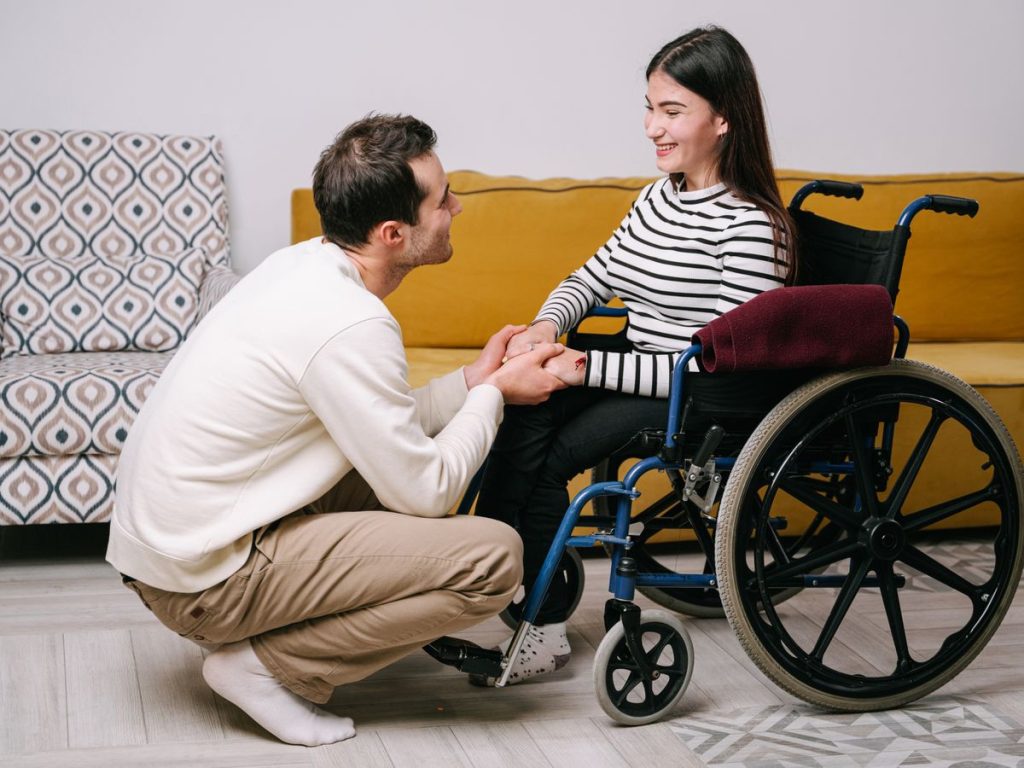 Man kneeling to hold the hands of a woman in a wheelchair at home