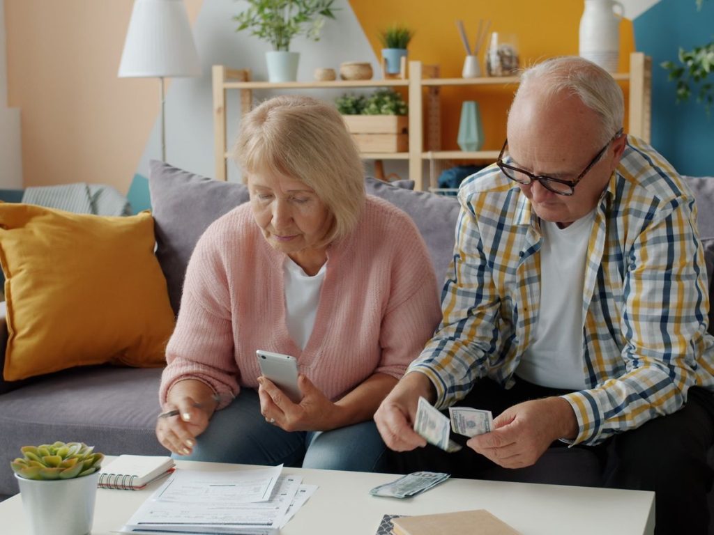 Elderly couple budgeting at home, seated on a cozy sofa, using smartphone and cash