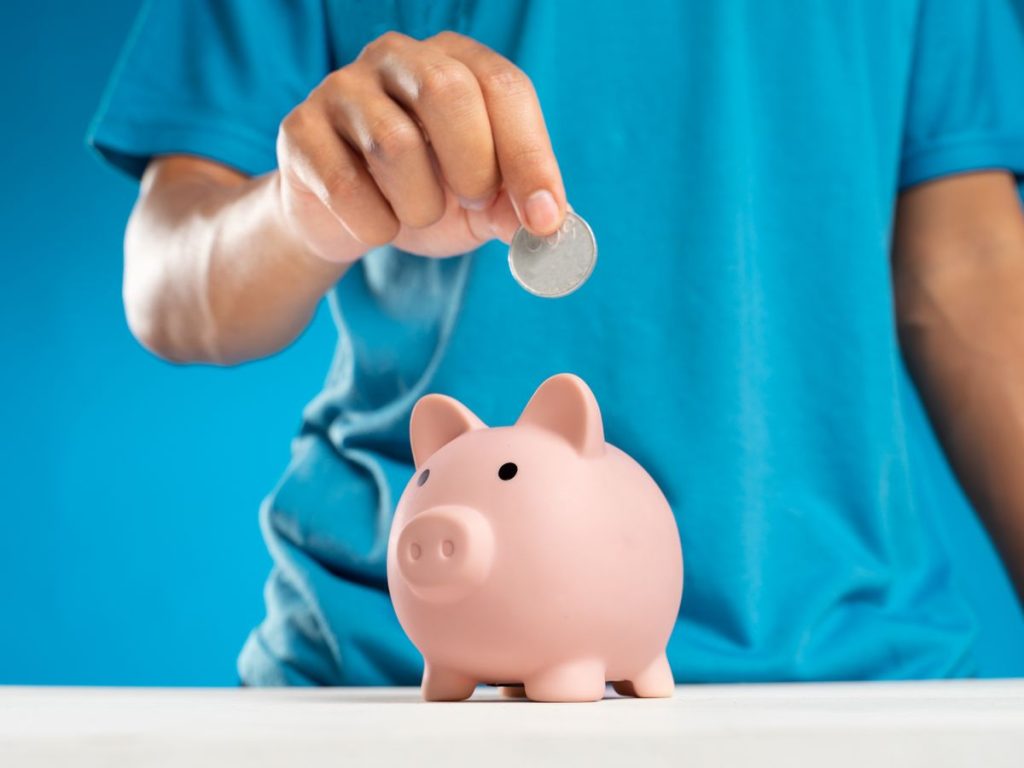 A close-up of an adult's hand dropping a coin into a piggy bank, symbolizing savings and investment