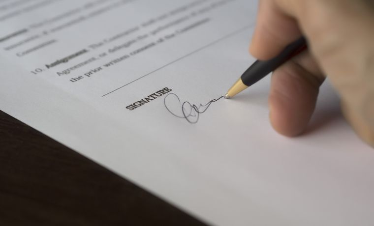 A hand signs a formal contract with a pen on a wooden desk